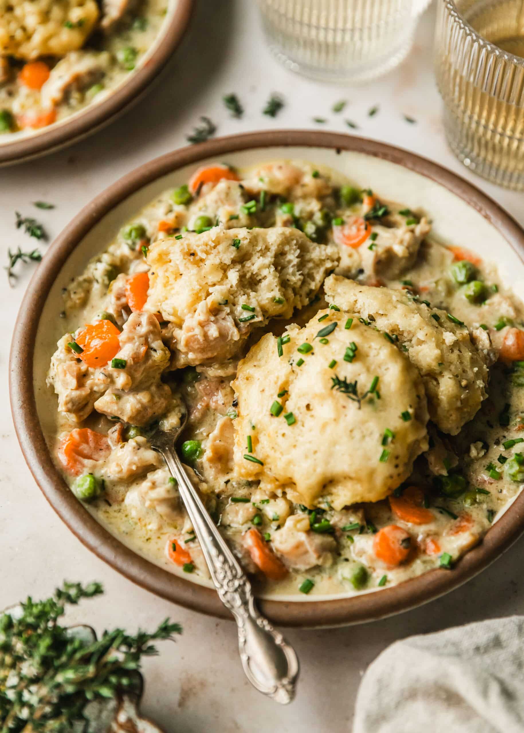 A brown plate of Dutch oven chicken and dumplings on a tan counter next to thyme, a beige linen, and glasses of white wine.
