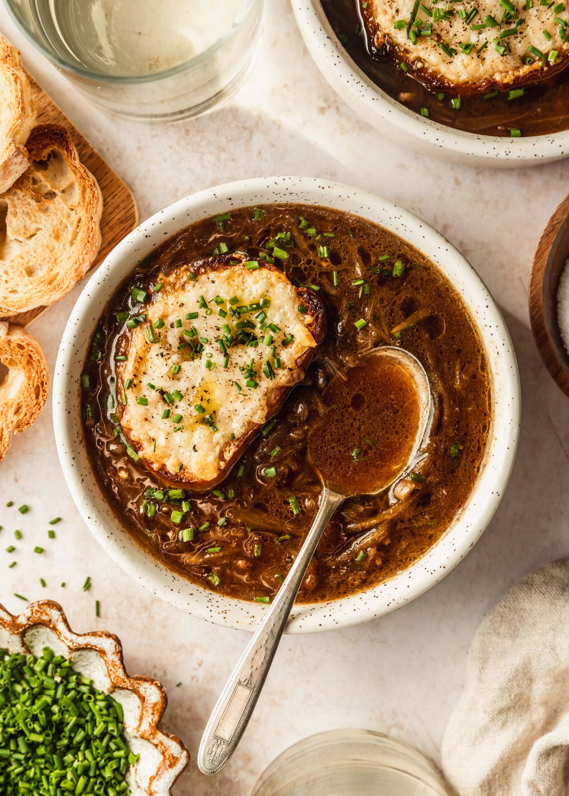 French shallot soup in a white bowl on a beige counter next to bread, a brown bowl of chives, and white wine.