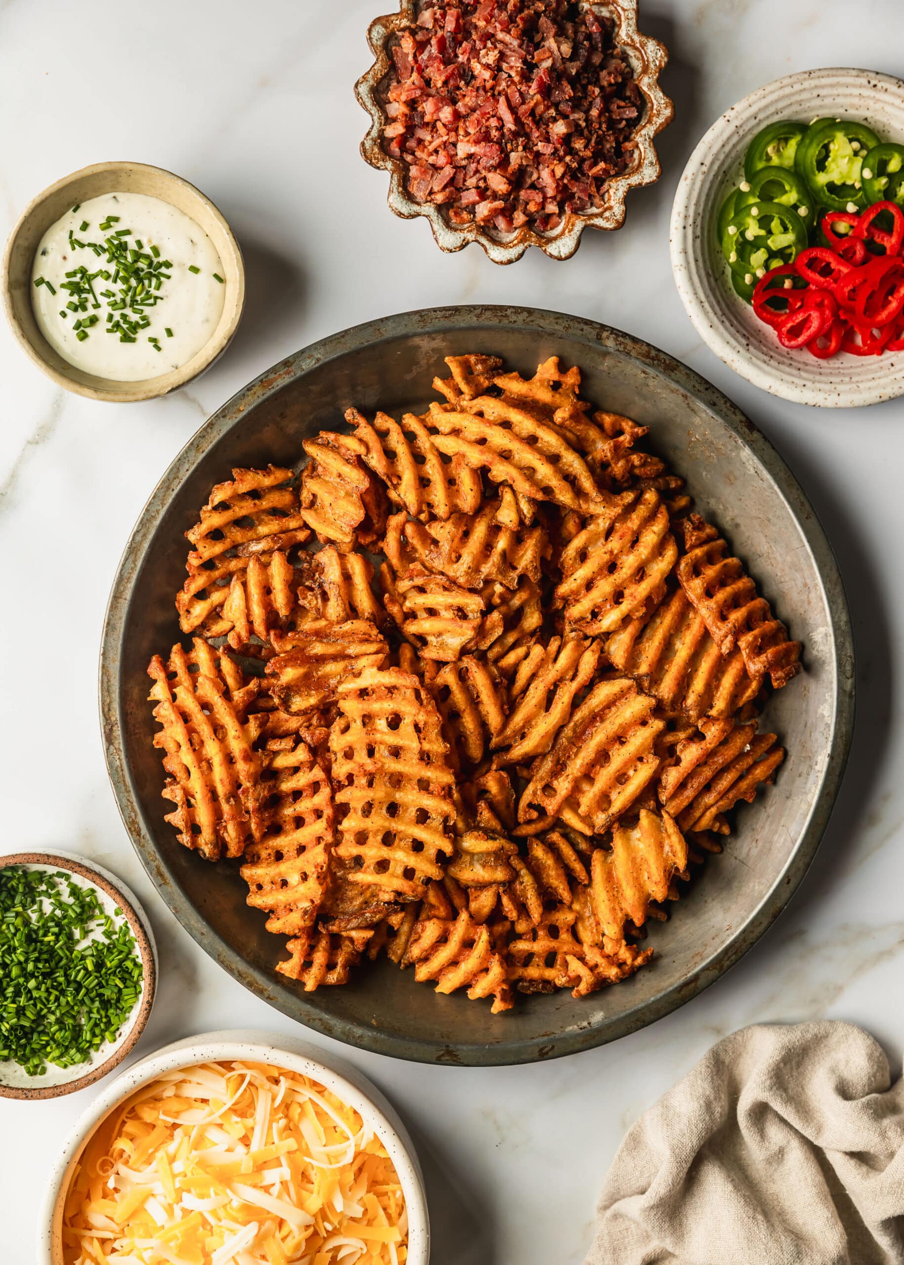 A tin of baked fries next to white and brown bowls of chives, cheese, peppers, bacon, and ranch on a white counter.