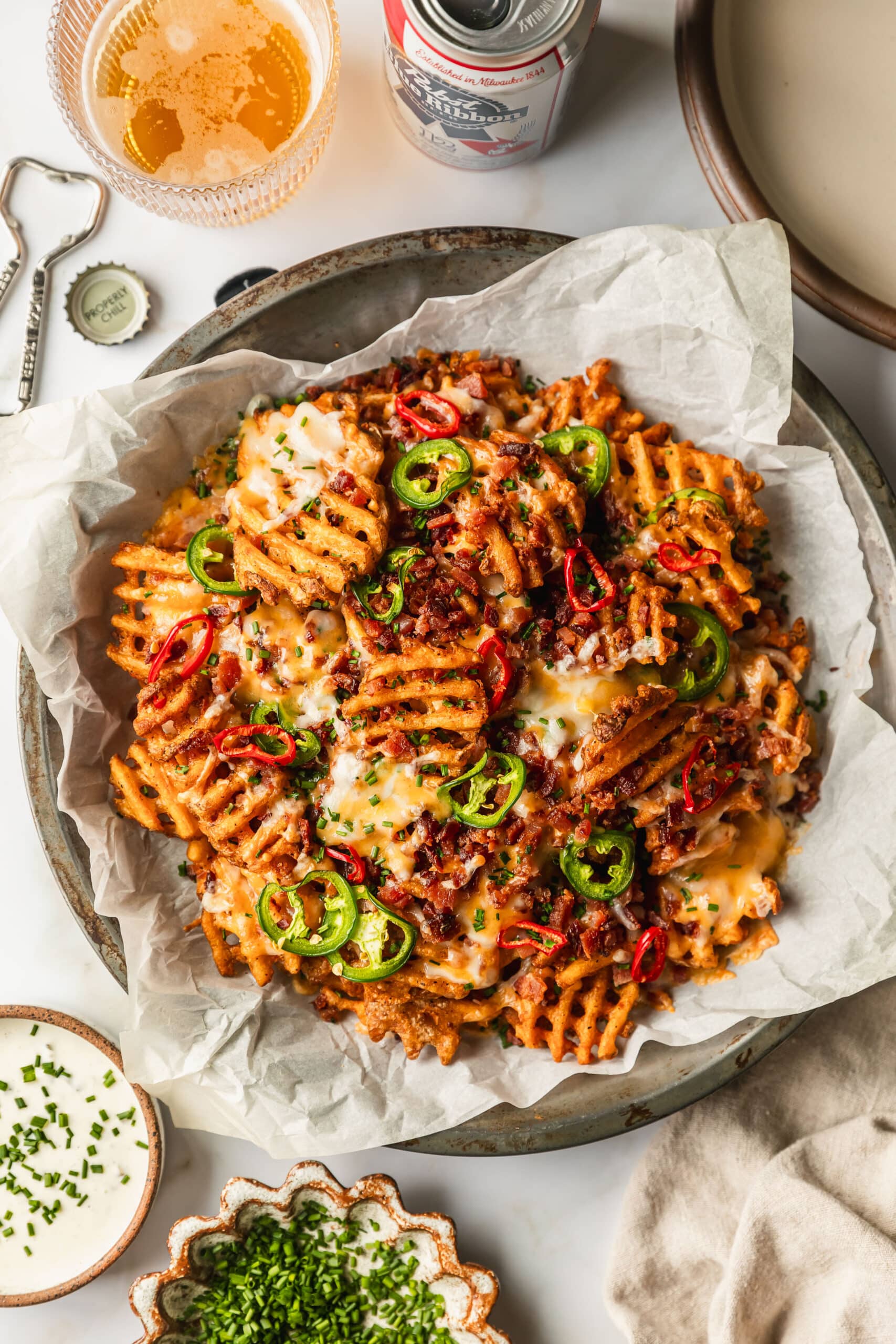 A metal pan of cheesy loaded waffle fries with bacon on a white counter next to beer and brown bowls of chives and ranch.