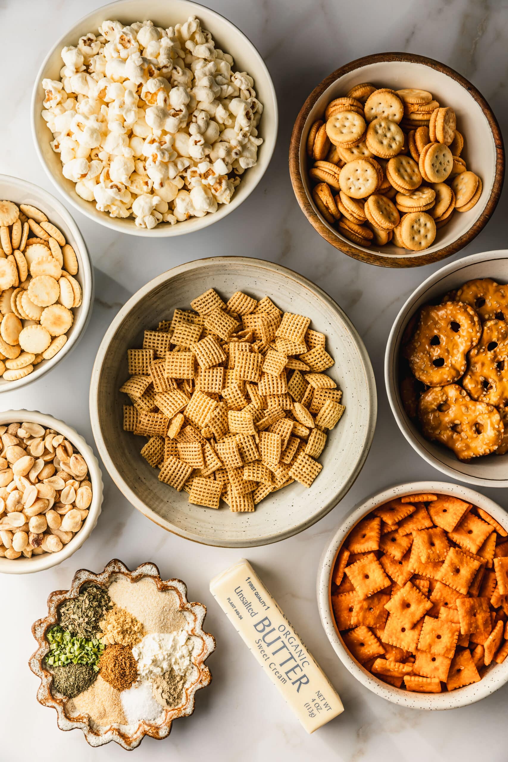 White and brown bowls of cereal, pretzels, Cheez Its, peanuts, spices, oyster crackers, sandwich crackers, popcorn, and butter on a white counter.