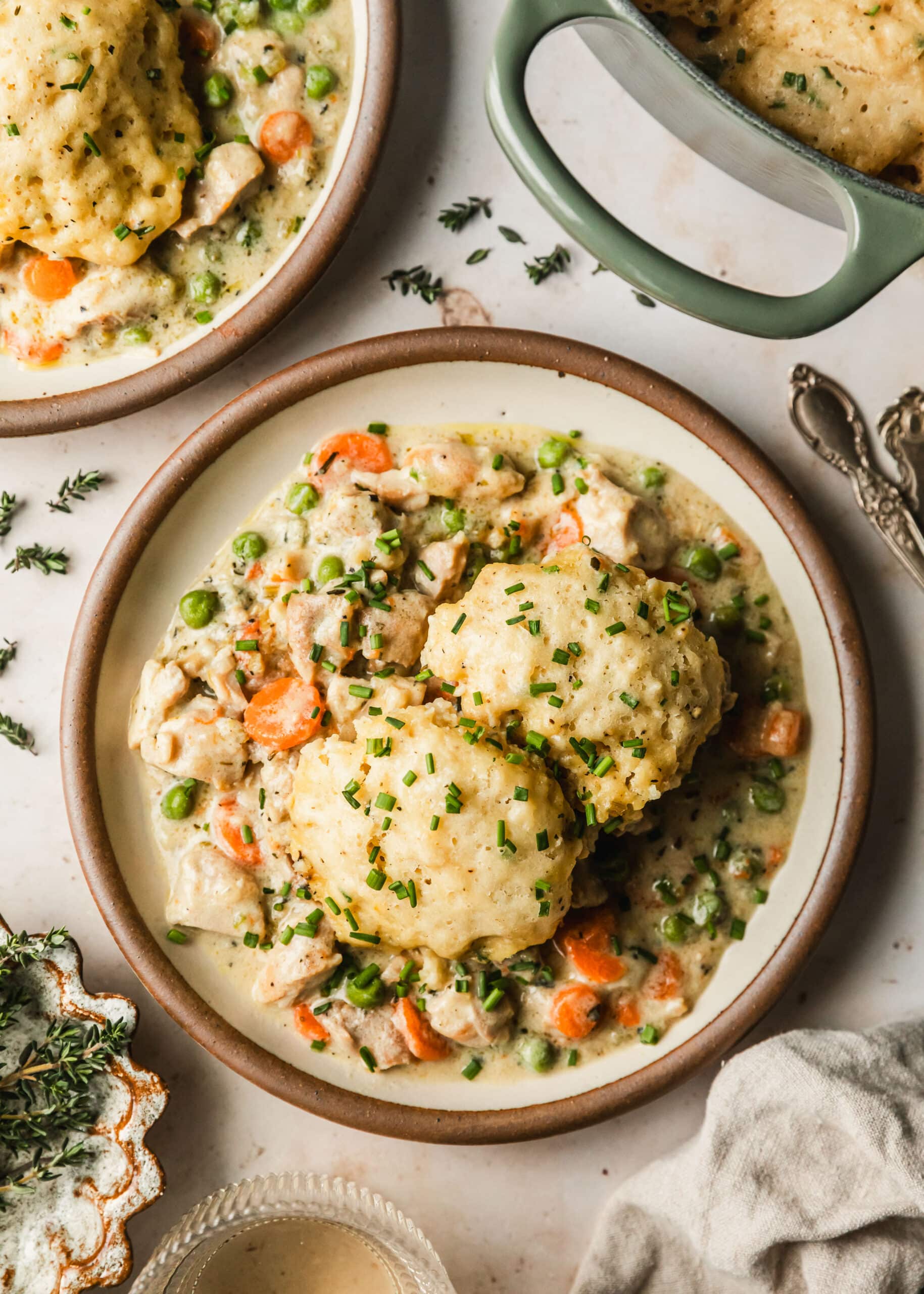Two white plates and a Dutch oven with chicken and dumplings on a beige counter next to thyme, a beige linen, and white wine.