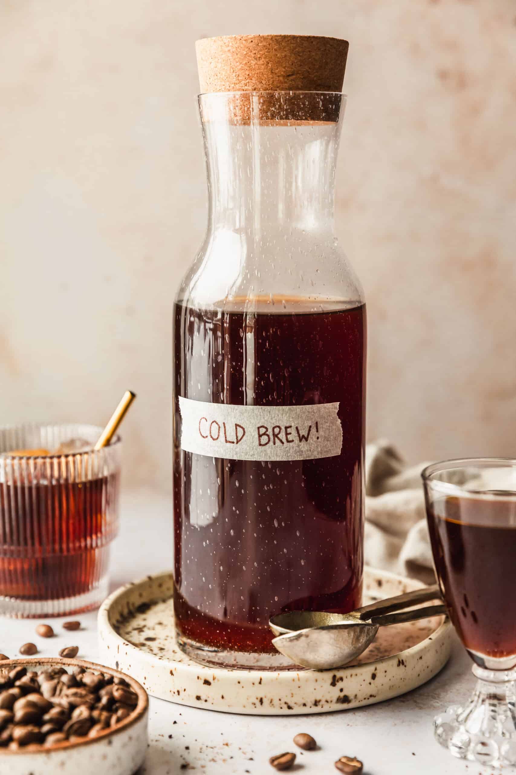 A carafe with cold brew on a beige background next to a white bowl of coffee beans, glass of cold brew, and glass of cold brew coffee concentrate.