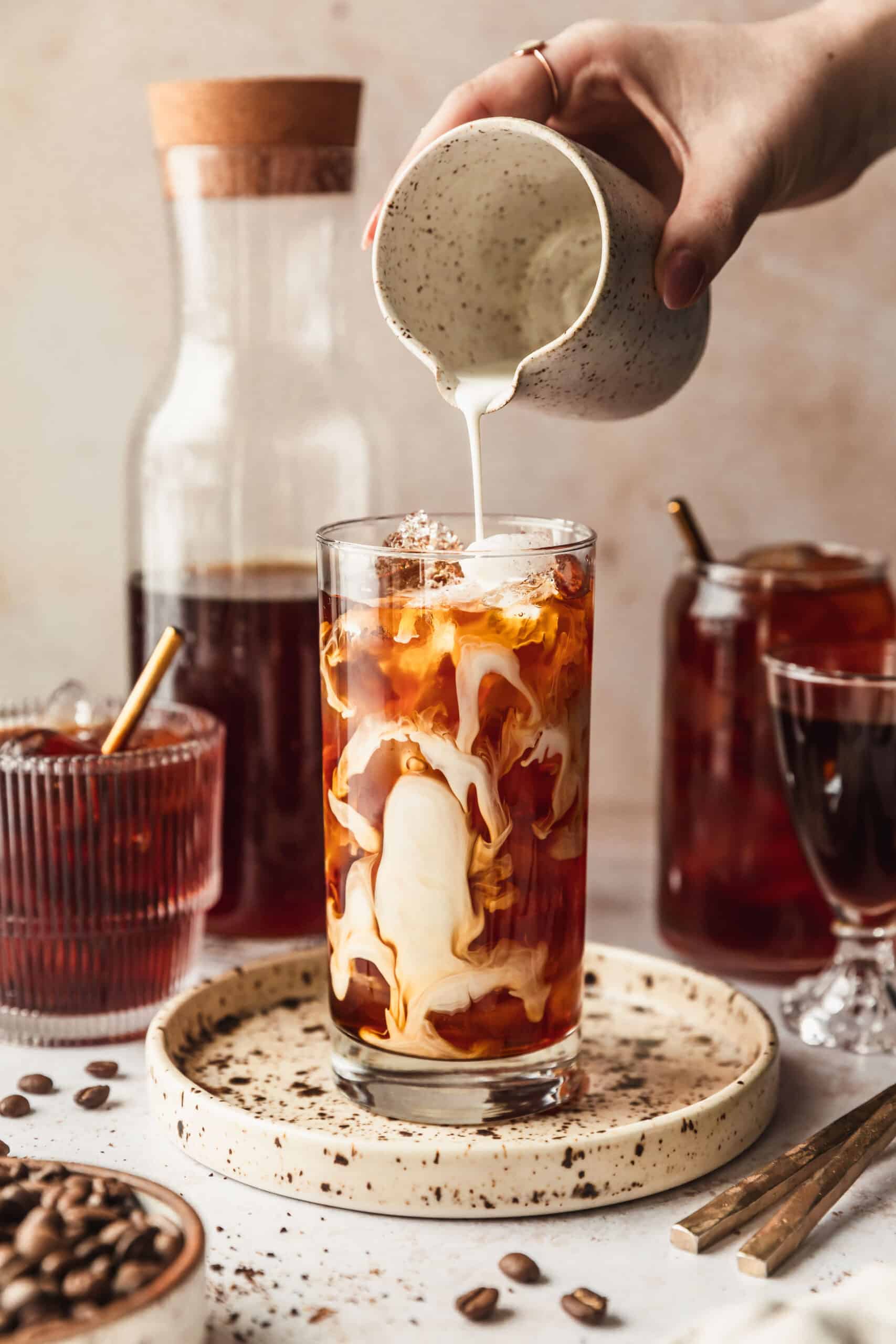 A hand pouring a a white cup of half-and-half into a cup of cold brew coffee concentrate on a beige counter next to a carafe of cold brew and glasses of cold brew.