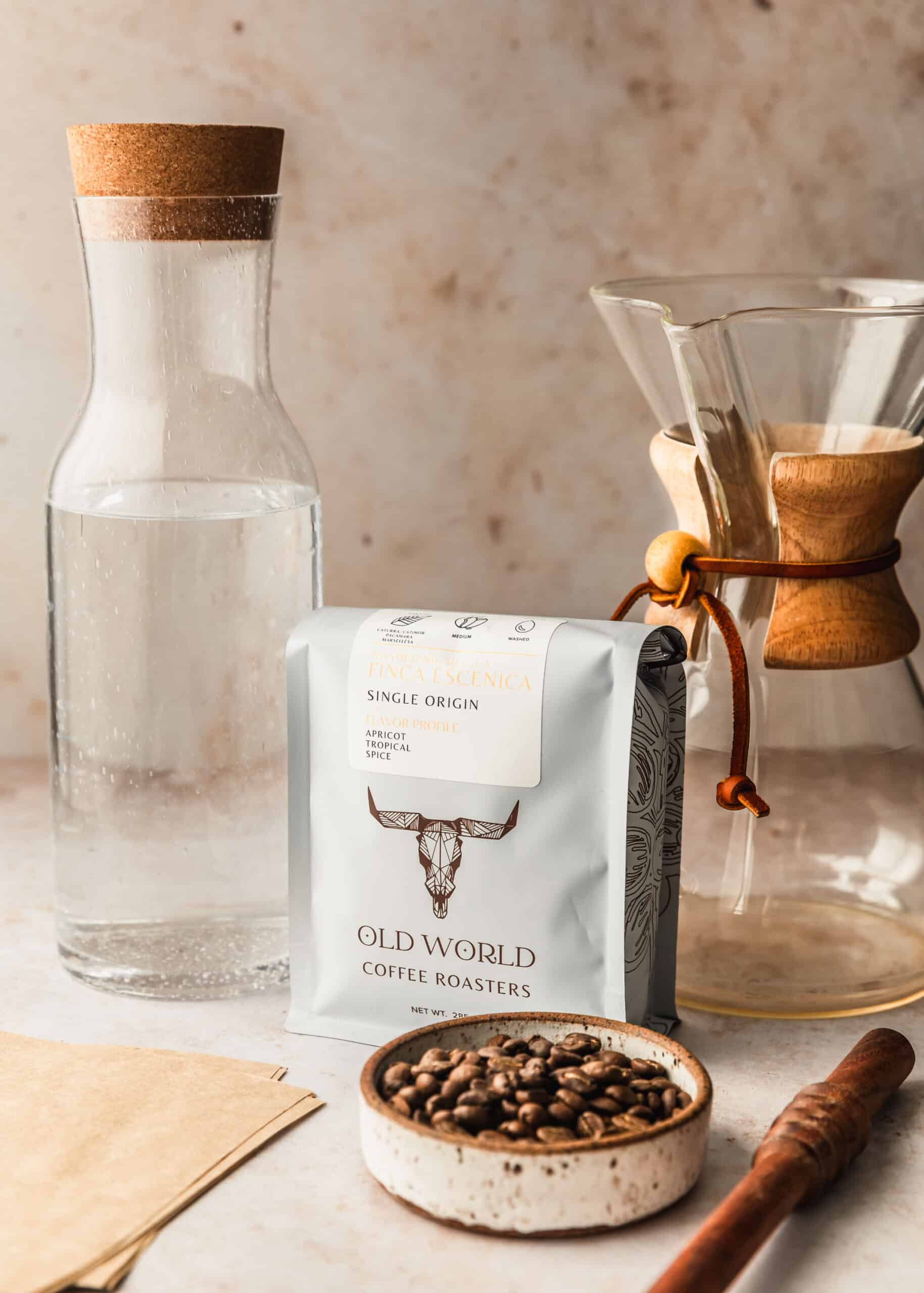 A bag of coffee beans on a beige counter next to a Chemex, pitcher of water, white bowl of coffee beans, and wood stirrer.
