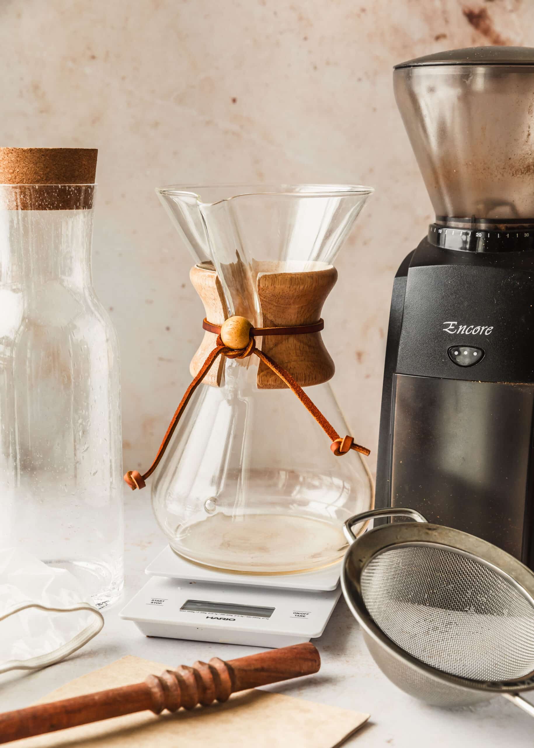 A Chemex, pitcher, grinder, scale, sieve, and wood spoon on a beige counter.