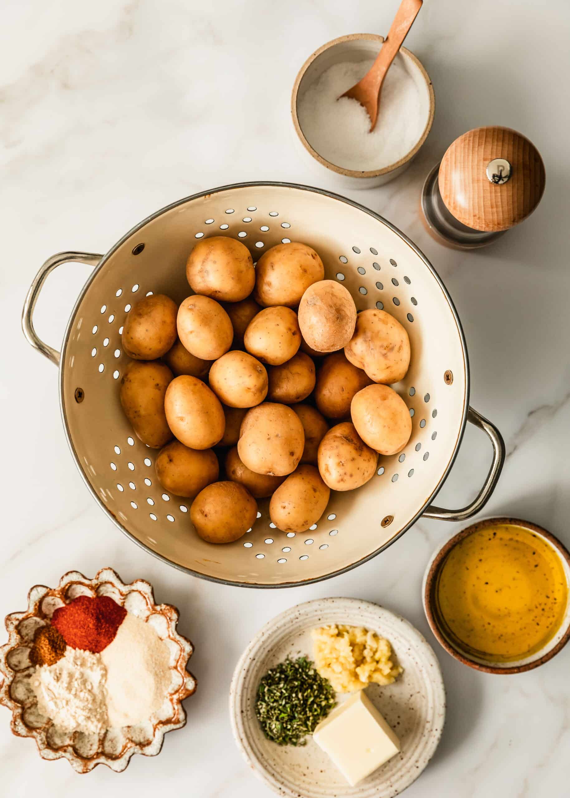 A white colander with mini Yukon potatoes next to a pepper grinder and white and brown bowls of salt, spices, garlic, rosemary, butter, and oil.