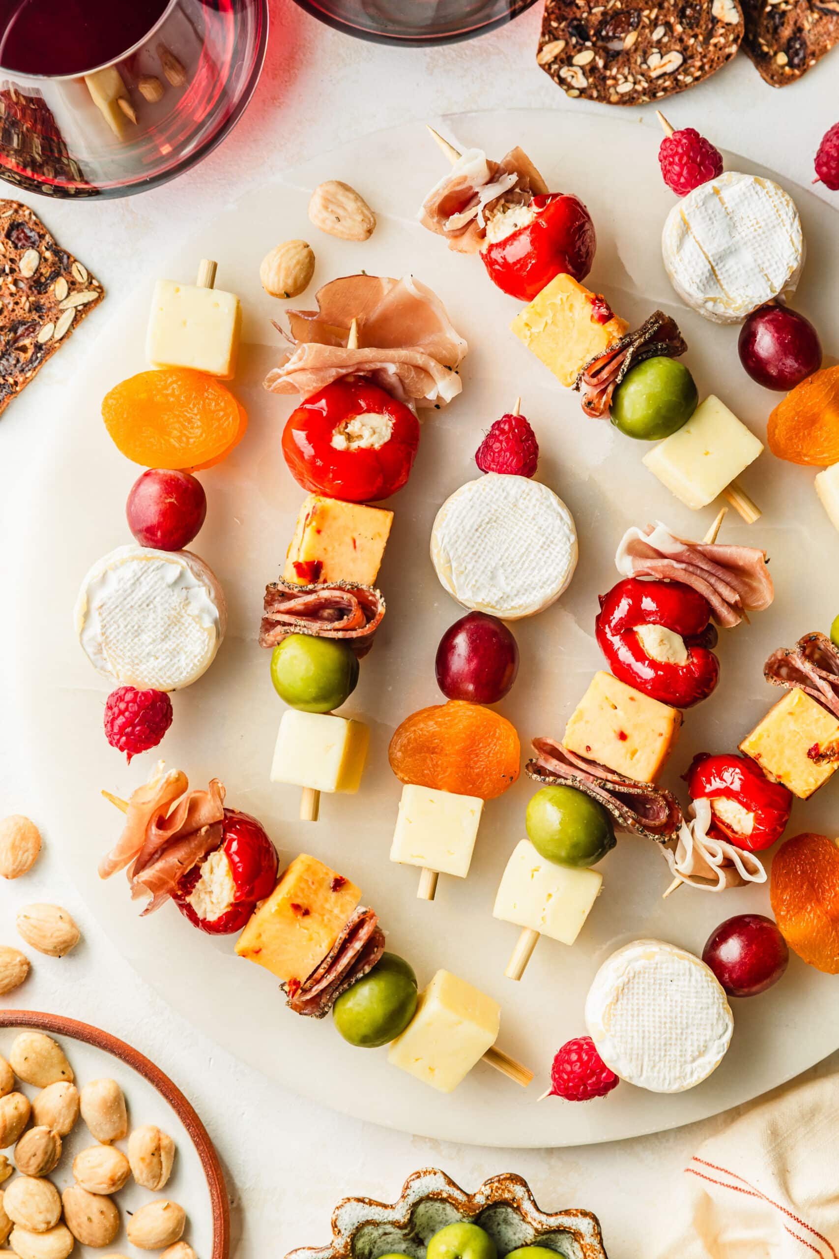 Charcuterie skewers on a white marble tray next to glasses of red wine, crackers, and brown paltes of olives and almonds on a white counter.