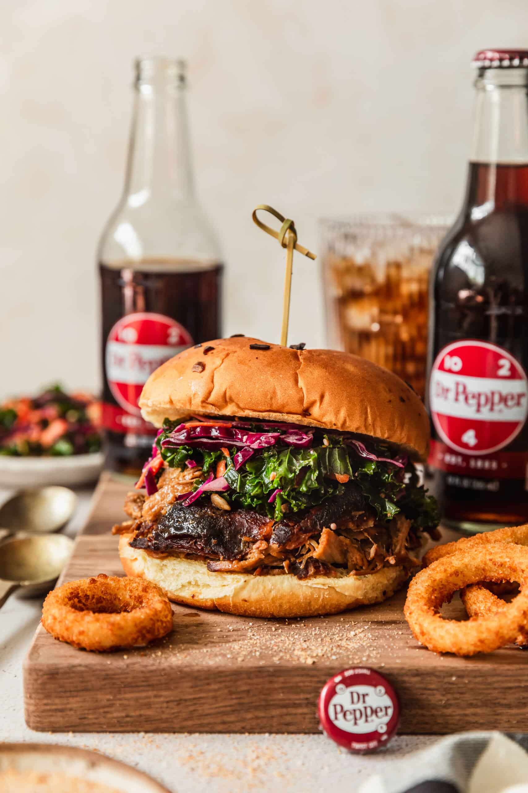 A sandwich with Dr. Pepper pulled pork on a wood board next to onion rings and bottles of Dr. Pepper on a tan counter next to a white bowl of slaw.