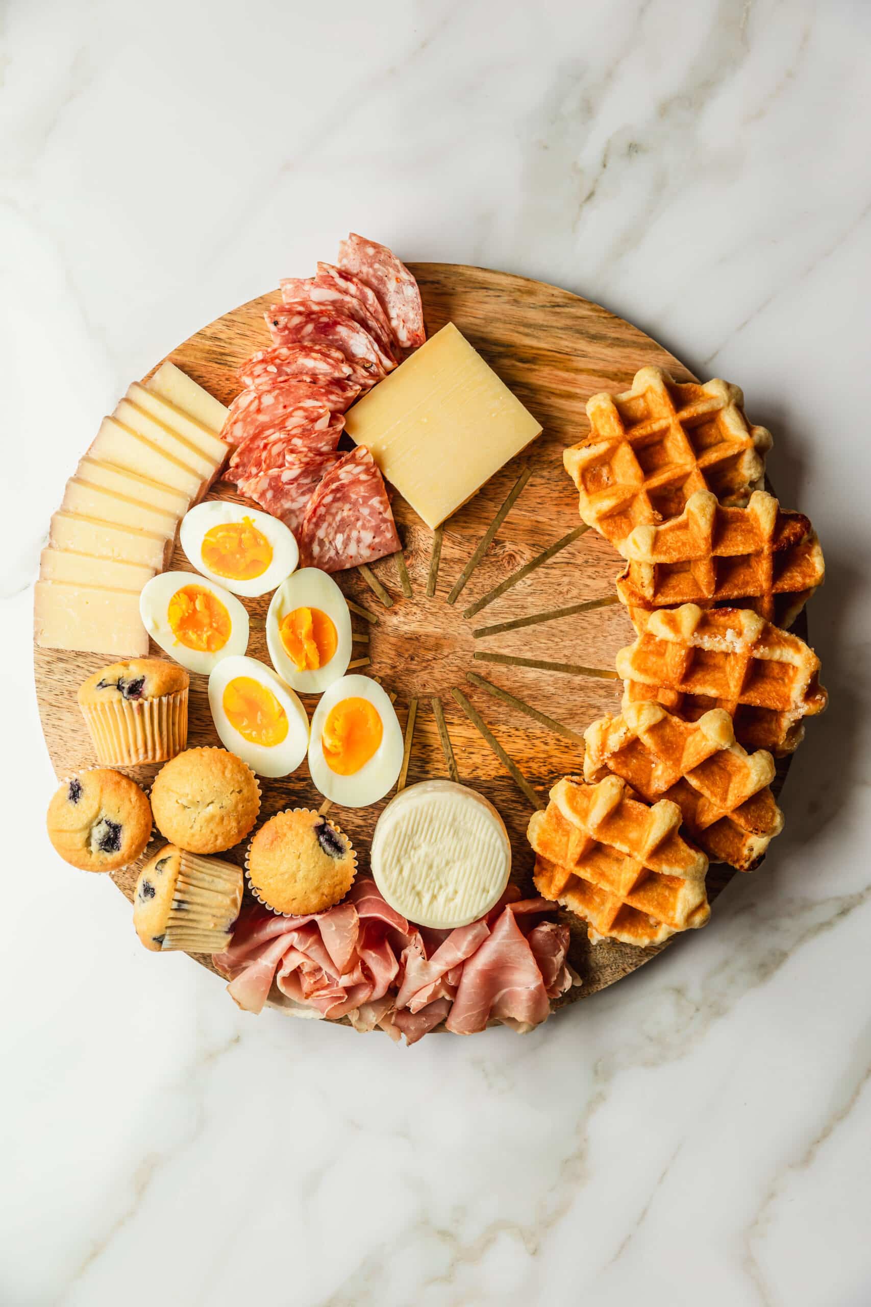 A wood board with meats, cheeses, eggs, waffles, and muffins on a marble counter.