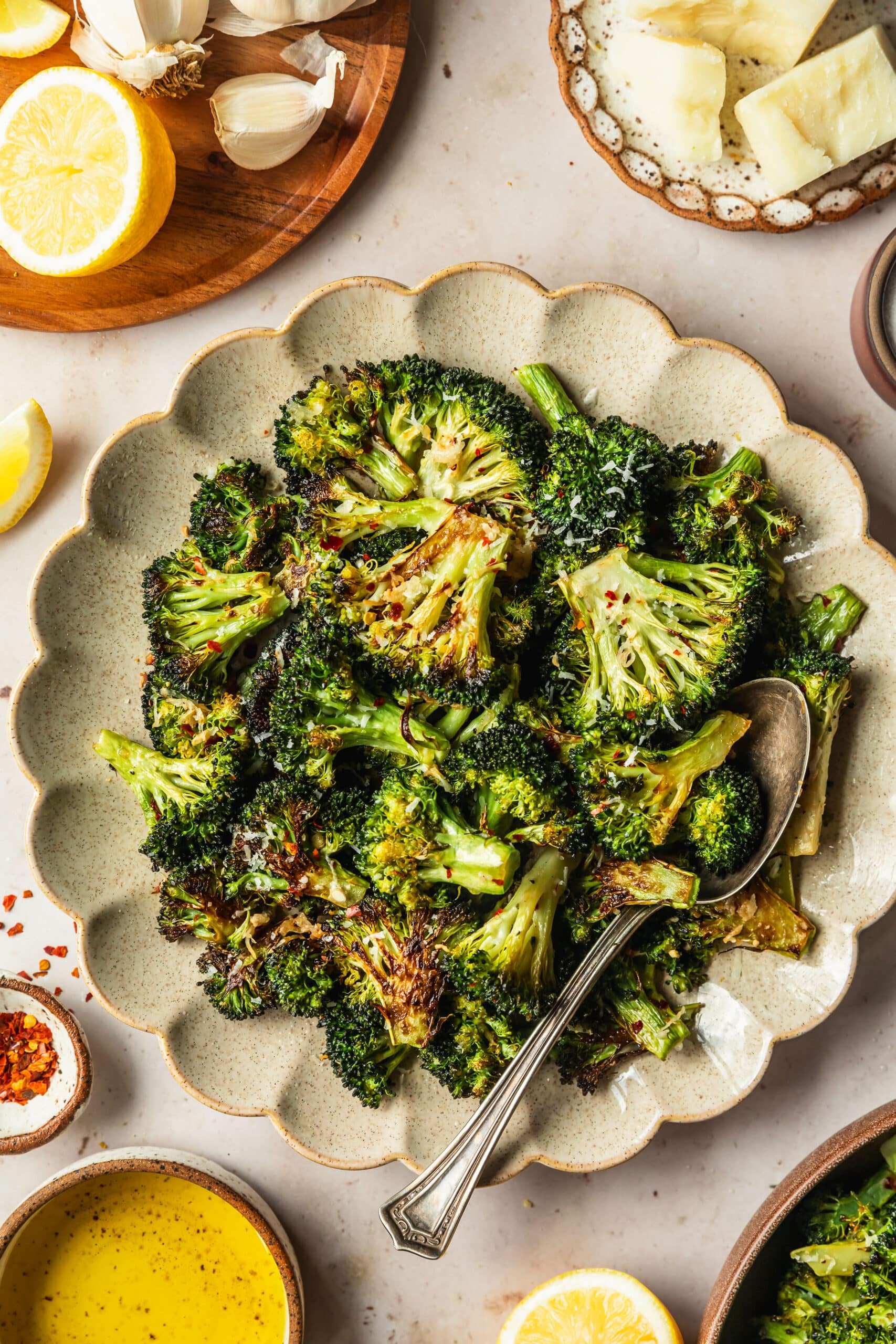 A tan plate of burnt broccoli on a tan counter next to white and brown bowls of parmesan, salt, lemons, oil, and pepper flakes.