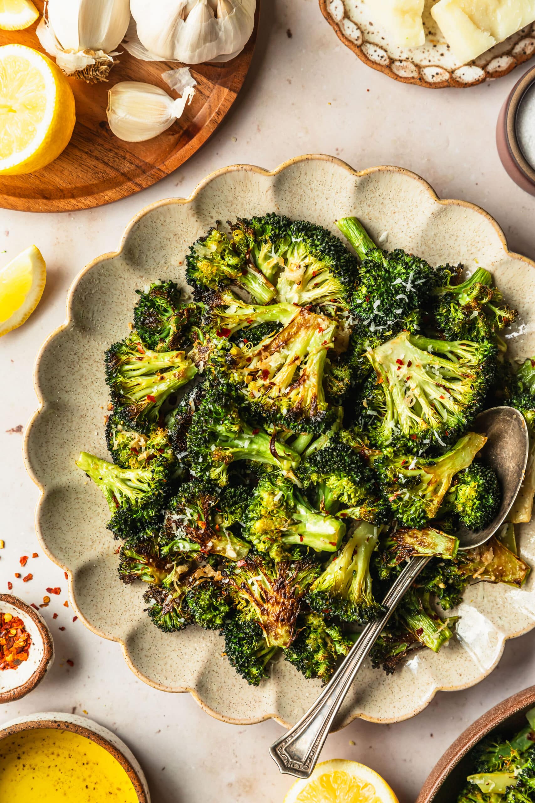 A cream-colored plate of charred broccoli on a tan counter next to white and brown bowls of parmesan, lemon, salt, red pepper flakes, oil, and more broccoli.