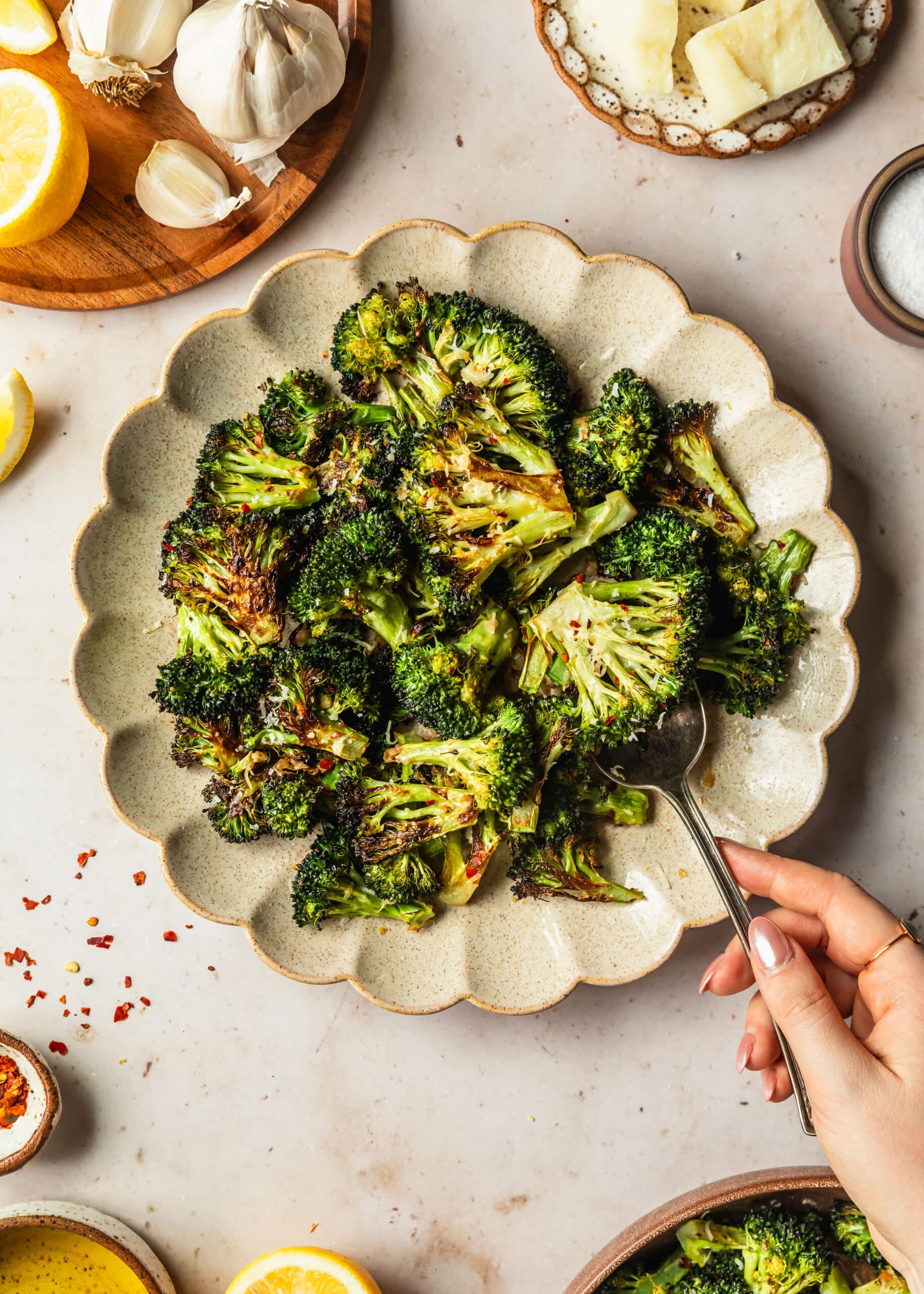 A hand spooning roasted broccolini off a cream platter on a tan counter next to brown bowls of parmesan, garlic, salt, red pepper flakes, and lemons.