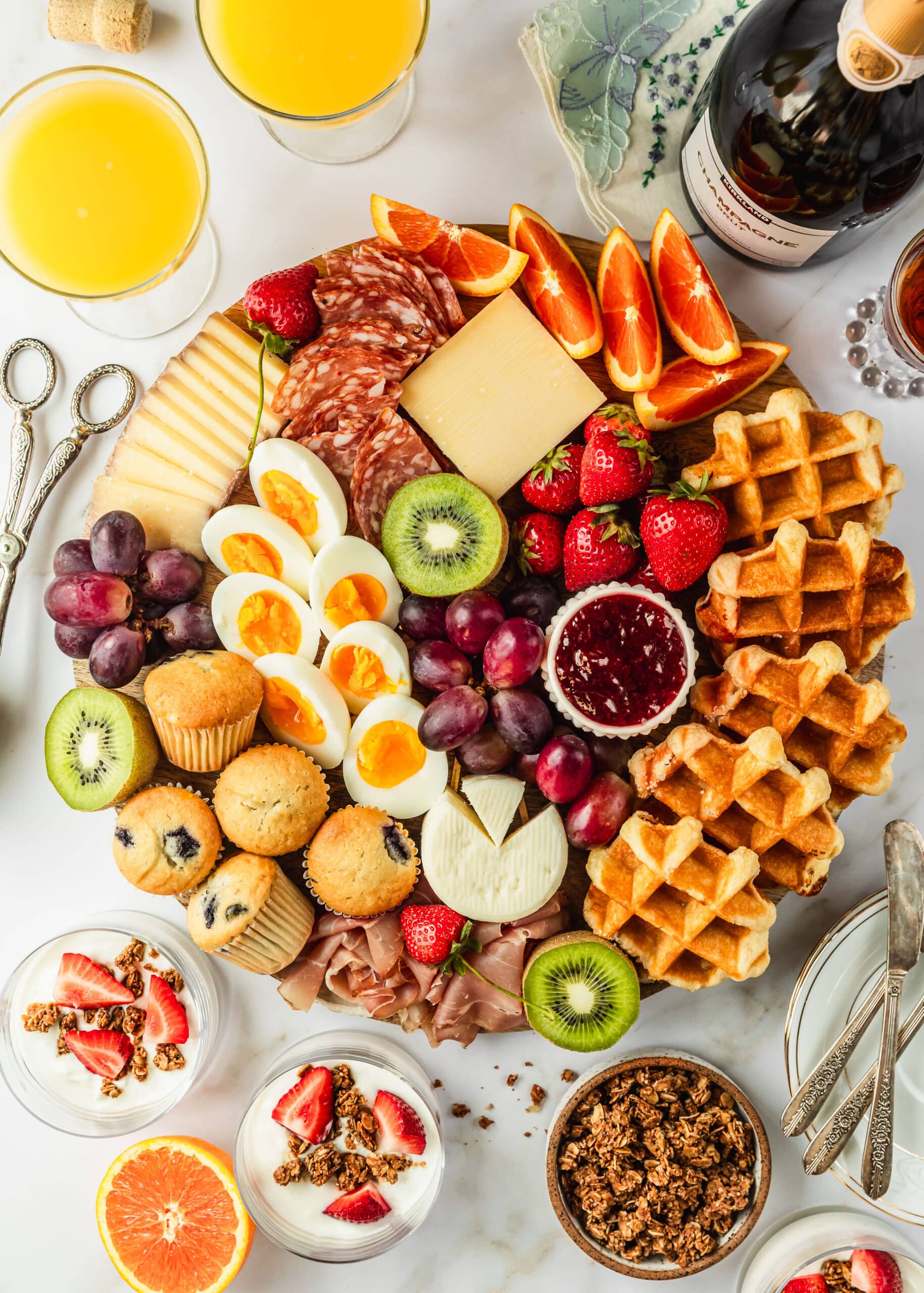 A brunch charcuterie board on a white counter next to mimosas, a bottle of champagne, and yogurt parfaits.