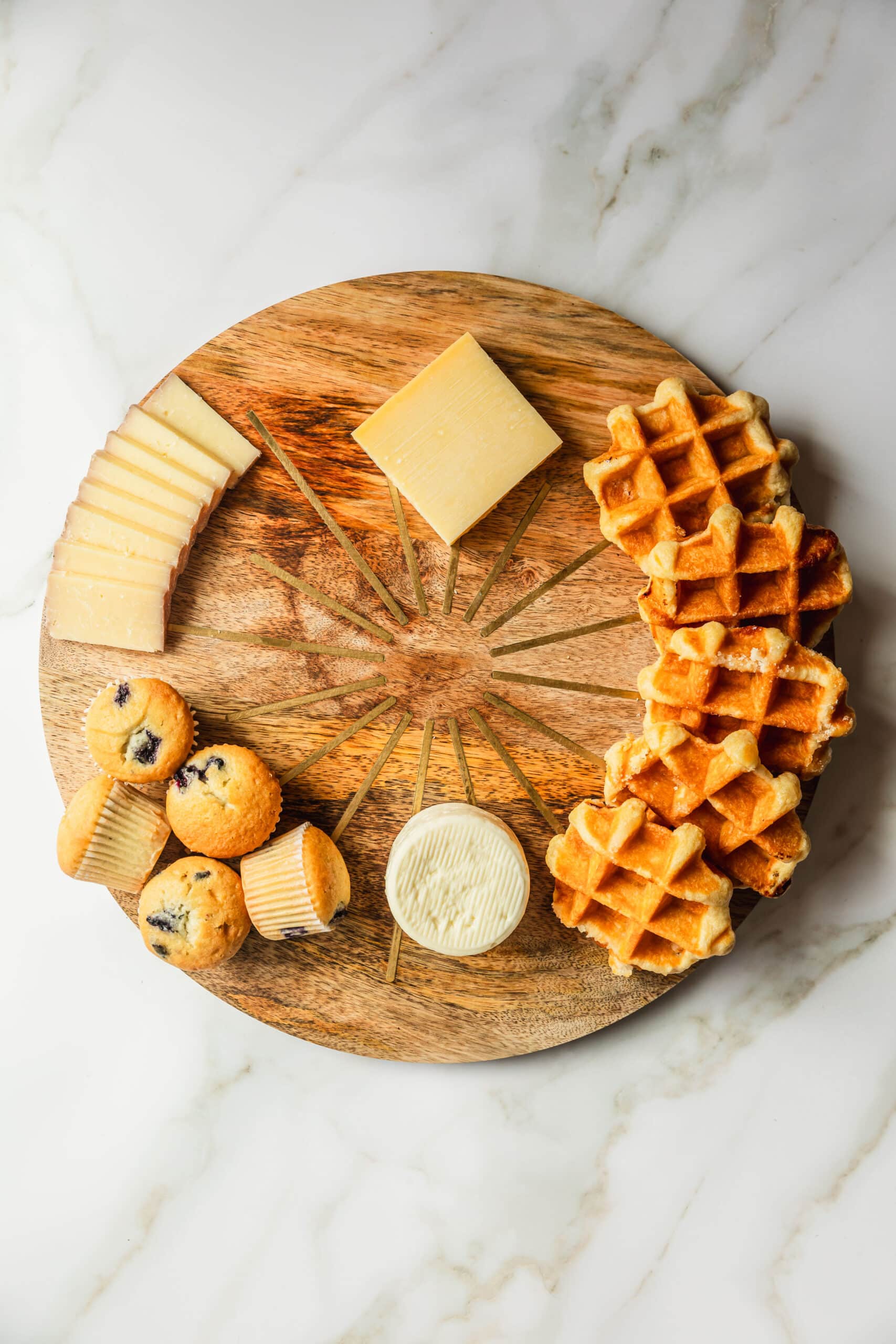 A board with cheeses, muffins, and waffles on a white counter.