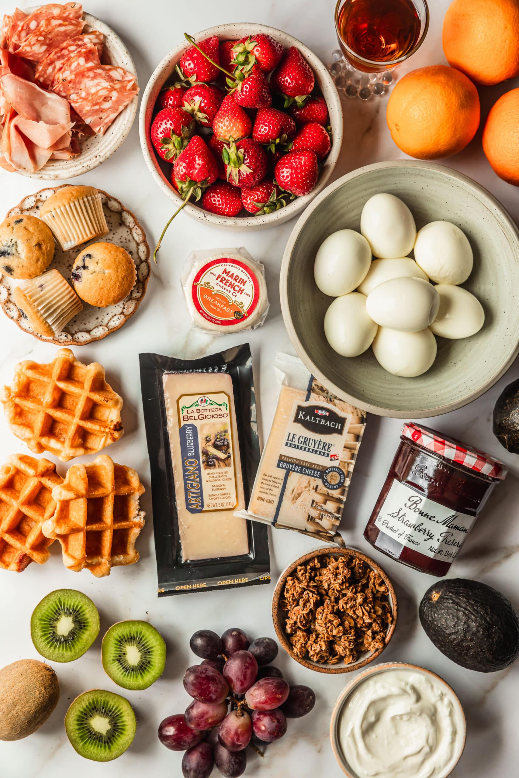 White bowls of meats, cheeses, berries, eggs, granola, yogurt, fruit, cheese, and waffles on a white counter.