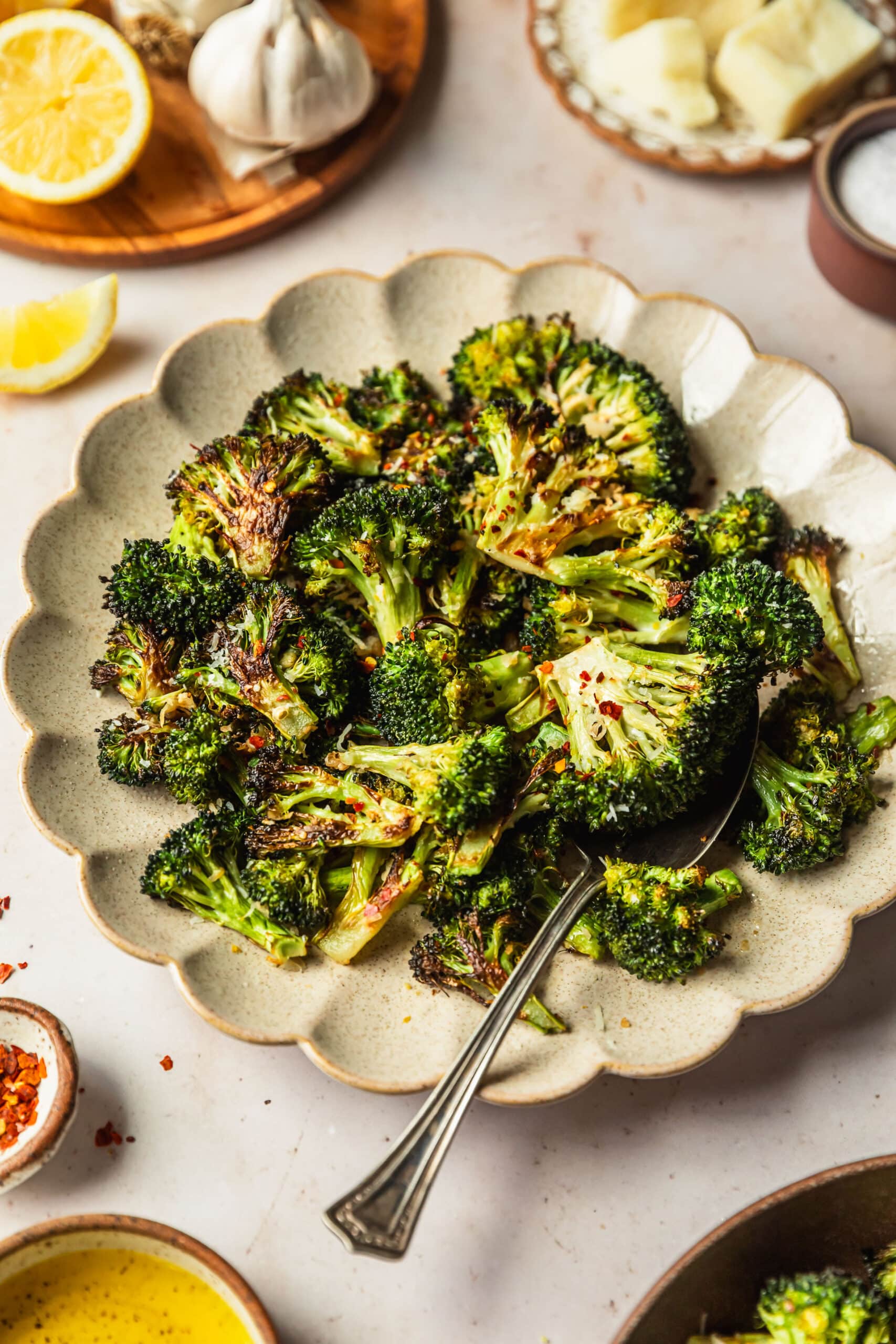Lemony charred broccoli with parmesan on a tan plate next to brown bowls of red pepper flakes, oil, salt, parmesan, and garlic with a tan background.