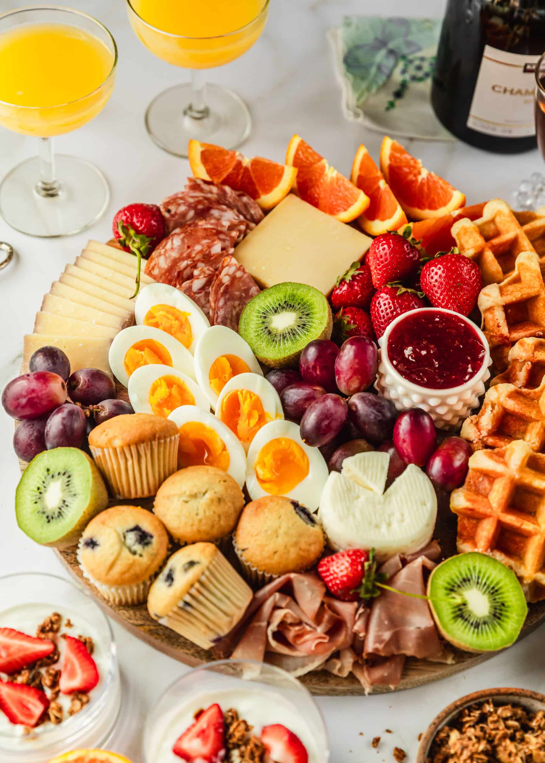 A brunch breakfast charcuterie board on a white counter next to yogurt parfaits, mimosas, and champagne.