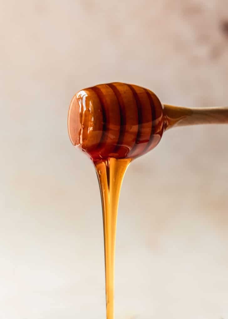 A honey dipper drizzling honey with a tan background.