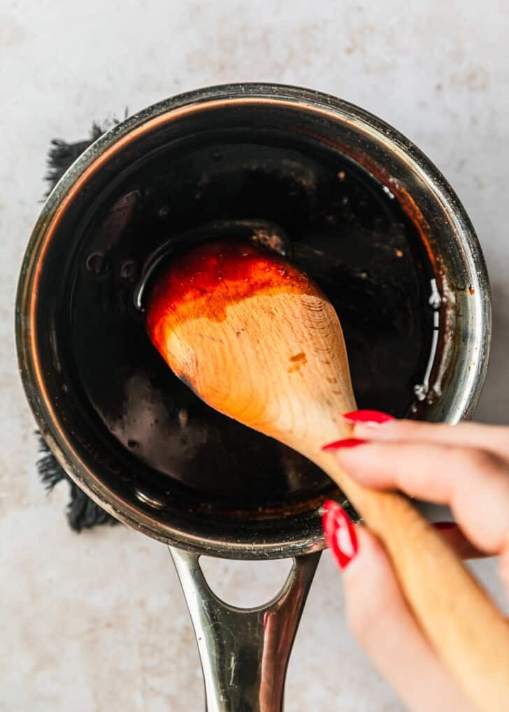 A hand using a wood spoon to stir a saucepan of balsamic vinegar on a tan counter.