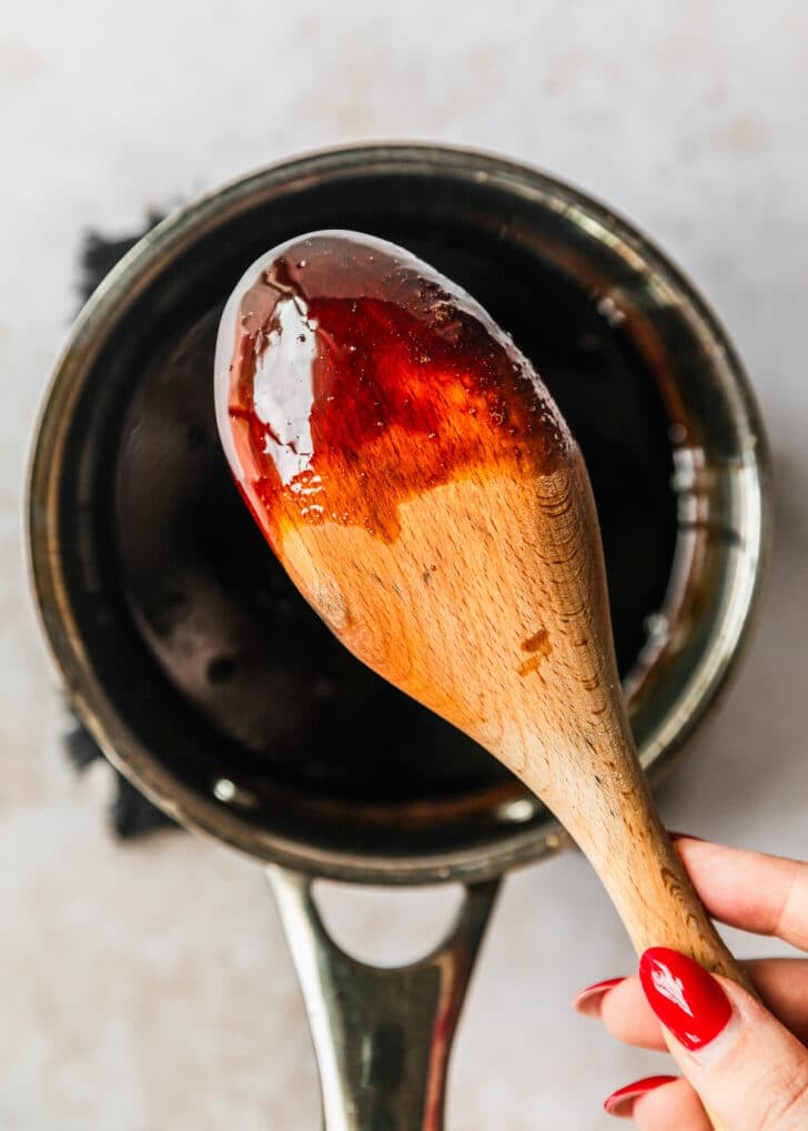 A hand holding a wood spoon covered in balsamic glaze over a saucepan of glaze.