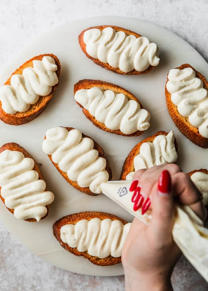 A hand using a piping bag to pipe whipped ricotta on a white platter of crostini.