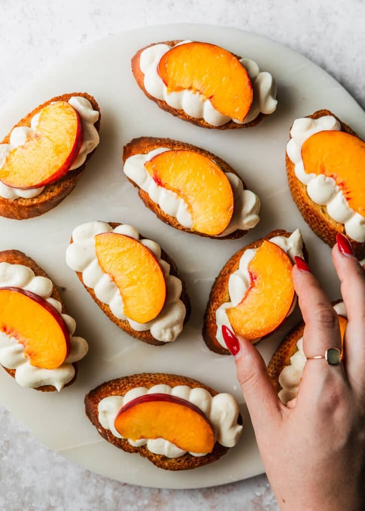 A hand placing peach slices on top of a white platter of crostini.