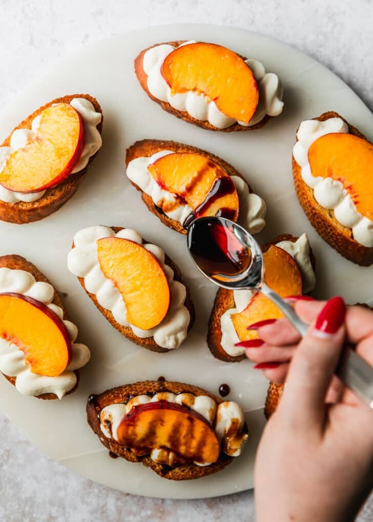 A hand using a spoon to drizzle balsamic glaze over peach crostini.