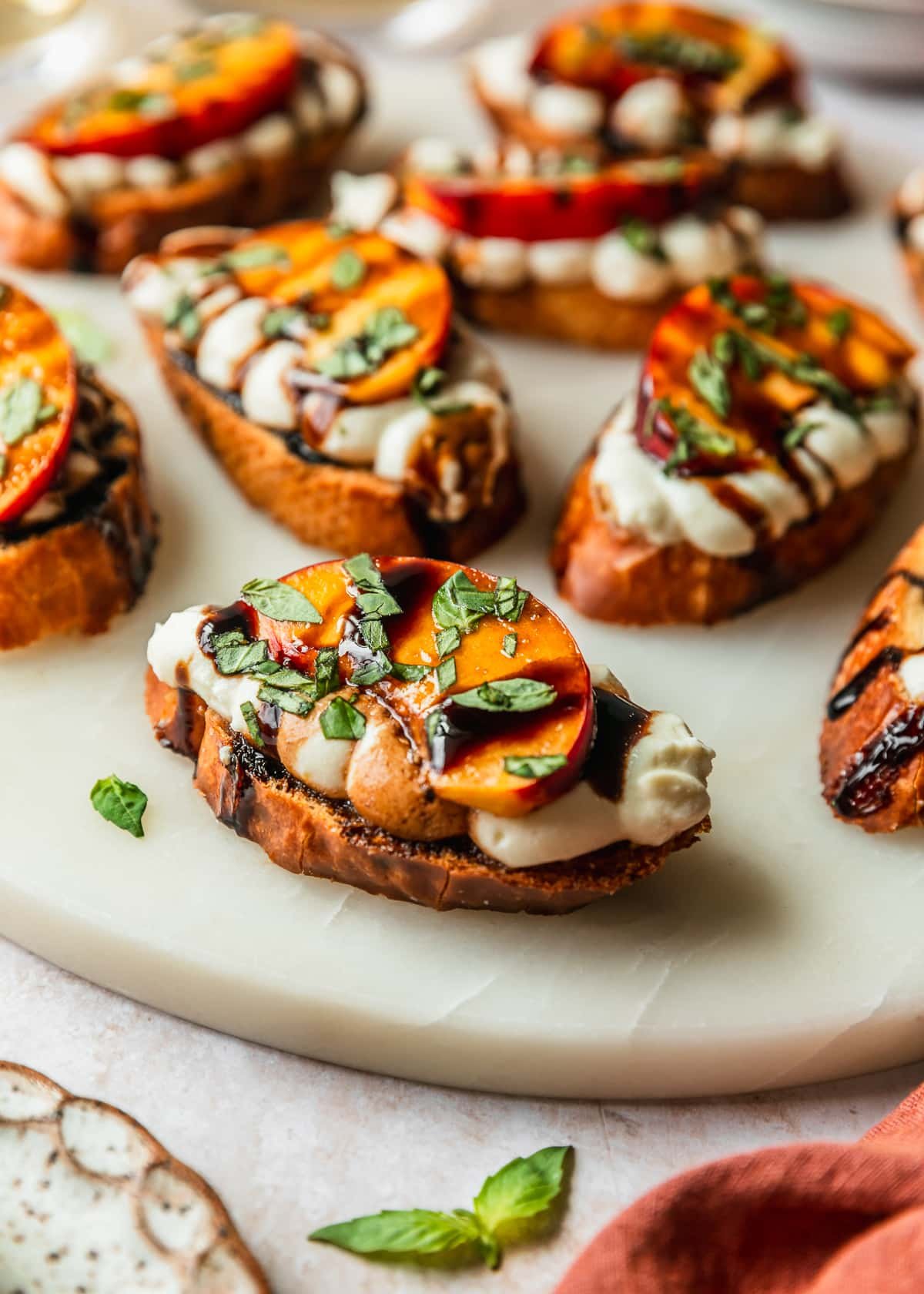 A white platter of peach and whipped ricotta crostini on a tan counter next to an orange linen, basil, and a white stoneware plate.