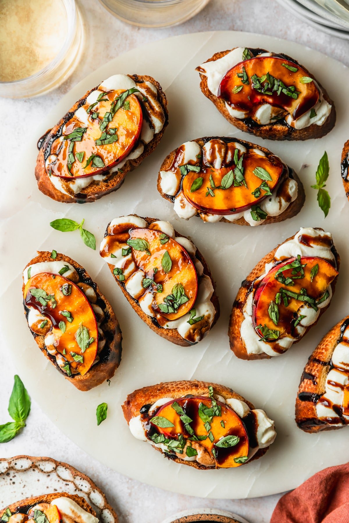 A marble platter of peach ricotta crostini on a marble counter next to white plates, glasses of white wine, and an orange linen.