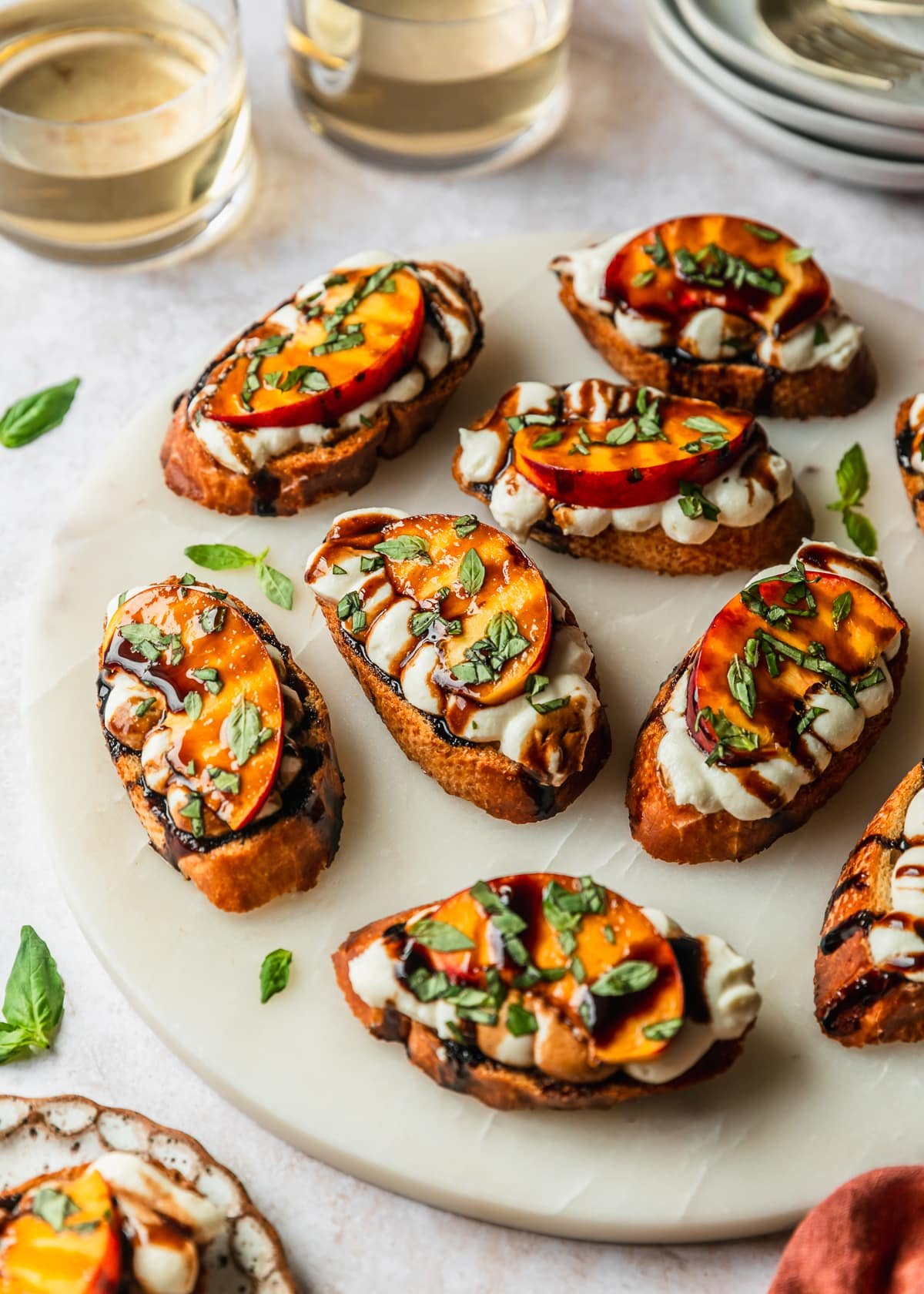 A marble platter of peach and whipped ricotta crostini on a tan counter next to white plates, glasses of white wine, and an orange napkin.