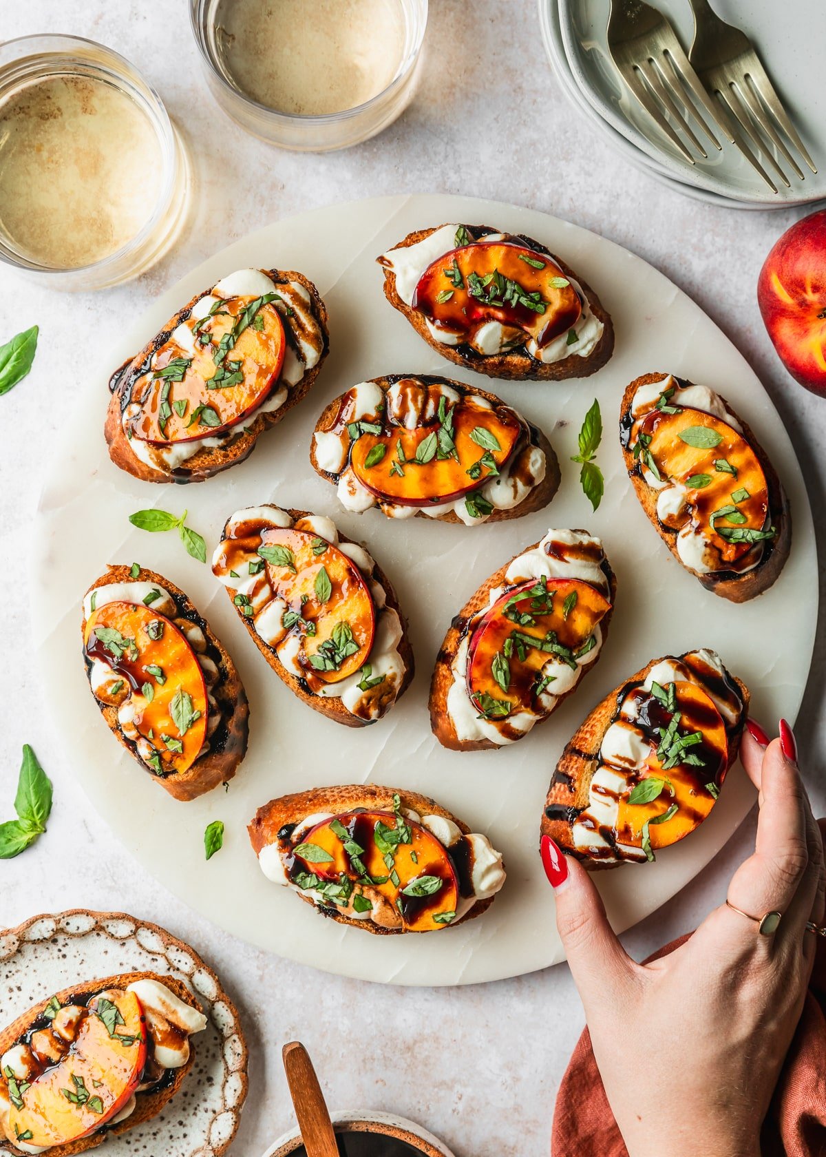 A hand reaching for a peach and whipped ricotta crostini on a white platter next to glasses of white wine, white plates, and an orange linen on a tan counter.