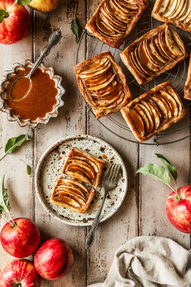 A tan plate and wire rack of mini caramel apple tarts on a wood table next to apples, a beige linen, and a brown bowl of caramel sauce.