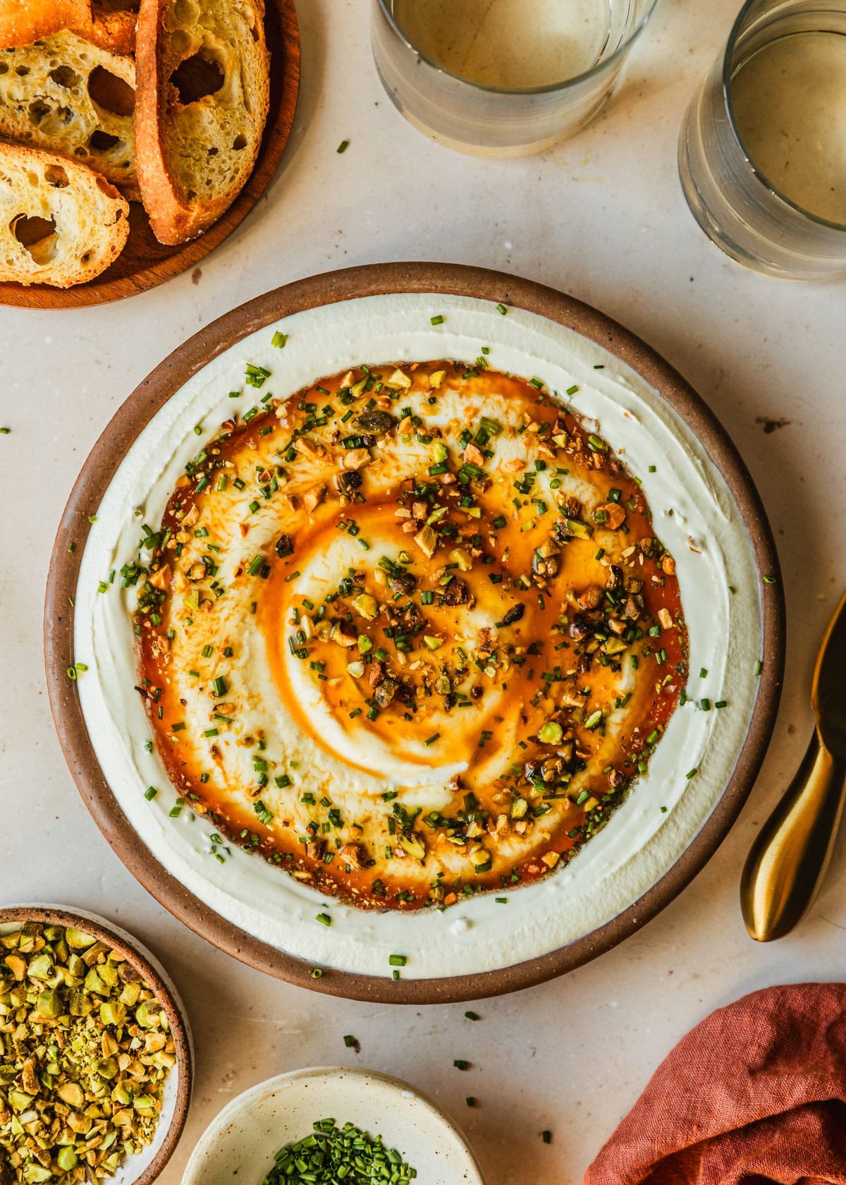 A brown bowl of whipped goat cheese with hot honey and pistachios on a tan counter next to white and brown bowls of pistachios, chives, and crackers, an orange linen, and glasses of white wine.