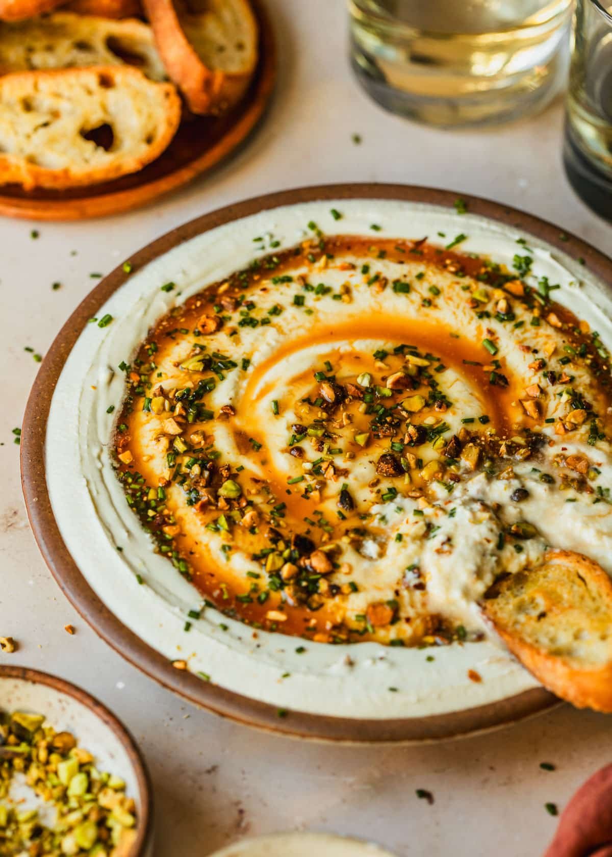 A stoneware bowl with whipped goat cheese, hot honey, and chives on a tan counter next to an orange linen, glasses of white wine, and a wood plate of crackers.