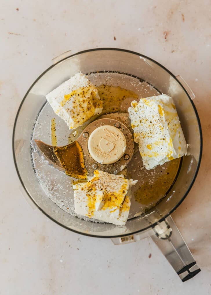 A food processor with cheese, olive oil, salt, and pepper on a tan counter.