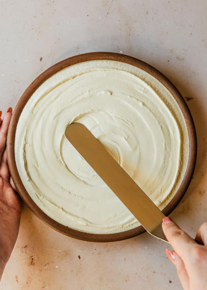 Hands spreading whipped goat cheese in a brown bowl on a tan counter.