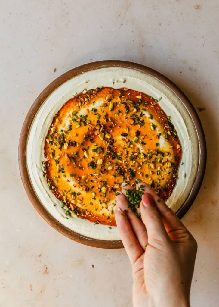 A hand sprinkling chives over a brown bowl of dip on a tan counter.