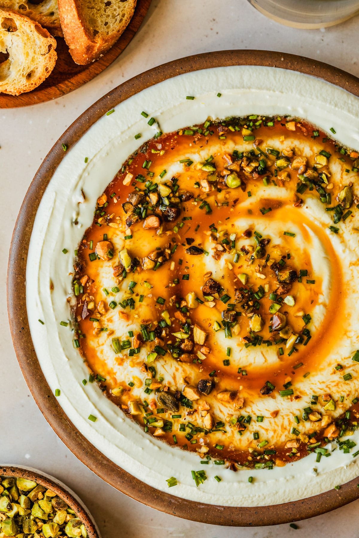 A brown bowl of whipped goat cheese with hot honey and pistachios on a tan counter next to brown bowls of pistachios and crackers.