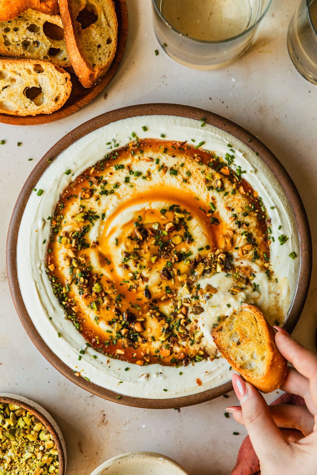 A hand dipping a cracker in a stoneware bowl of whipped goat cheese with hot honey and pistachios on a tan marble counter next to brown bowls of pistachios and crackers and glasses of white wine.