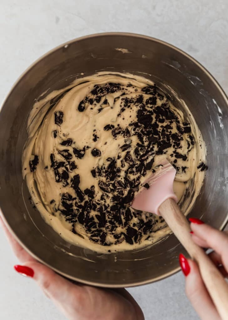 A hand using a rubber spatula to mix mini Oreo cheesecakes filling in a silver bowl.