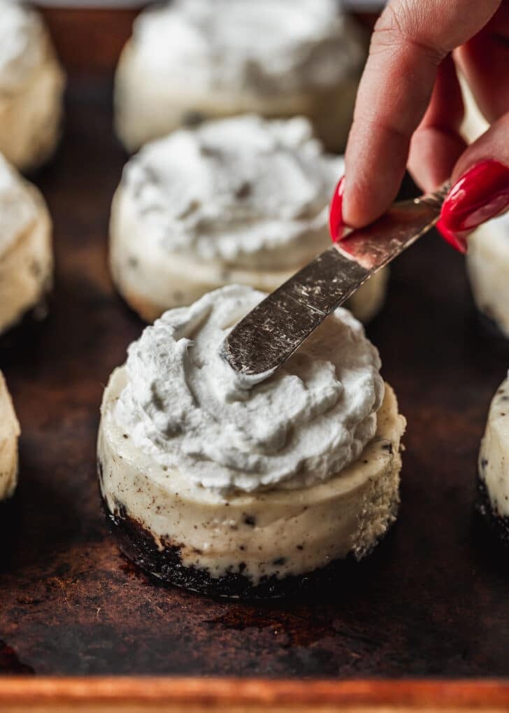 A hand using a knife to spread whipped cream on mini Oreo cheesecakes.