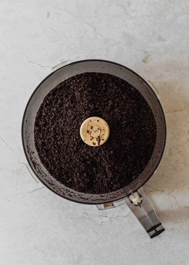 Chocolate cookie crumbs in a food processor on a marble counter.