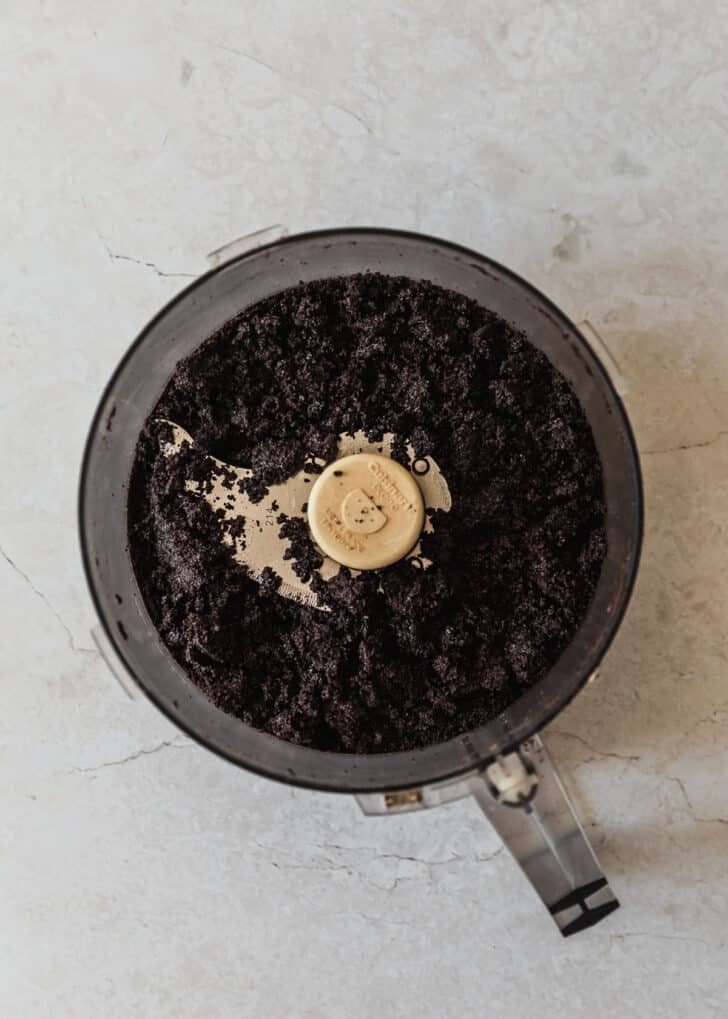 Chocolate cookie crust in a food processor on a white counter.