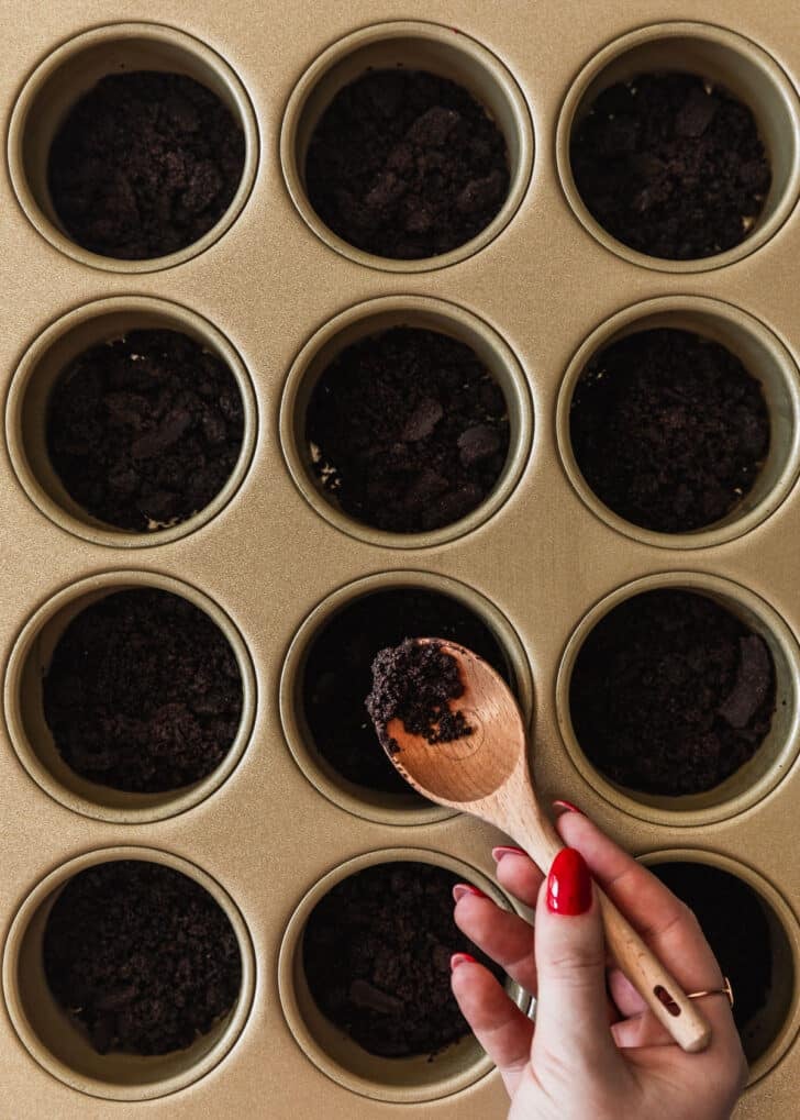 A hand spooning Oreo cookie crust into mini cheesecakes pans.