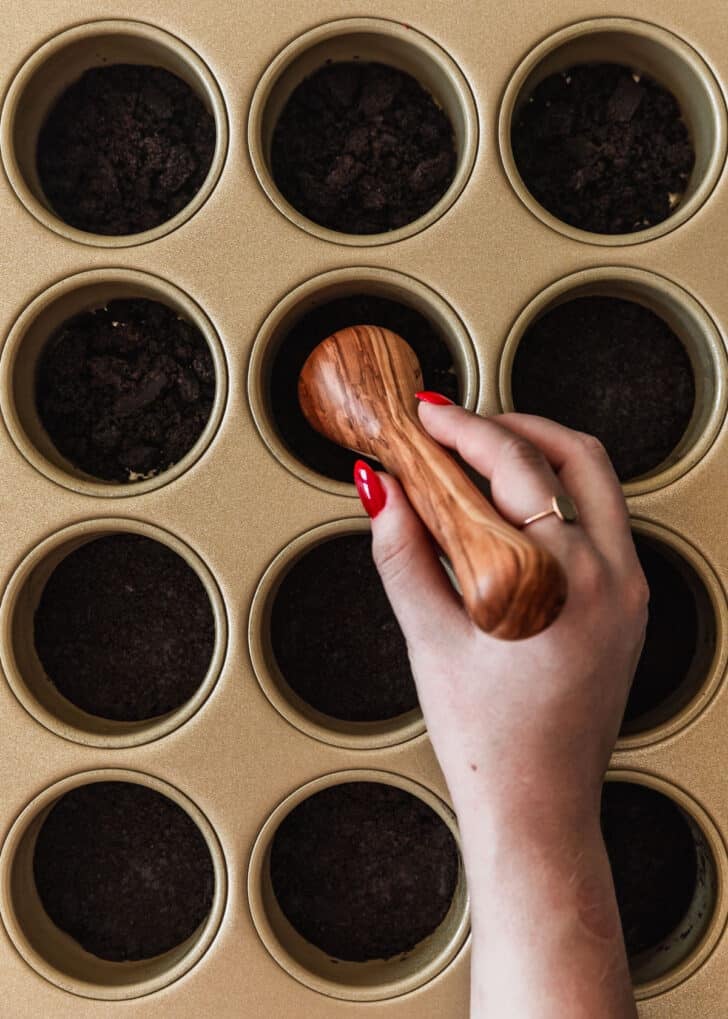 A hand using a pastry tamper to press chocolate crust into muffin pans.