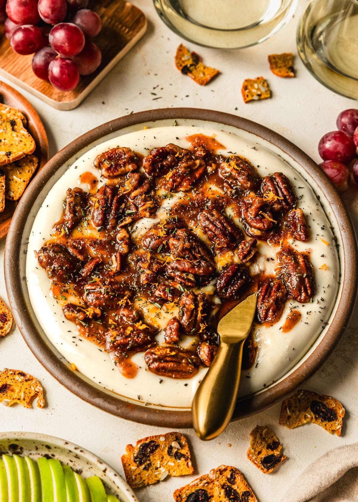 A brown plate of whipped brie cheese with sticky pecans on a beige counter next to glasses of white wine and brown plates of grapes, sliced green apples, and crackers.