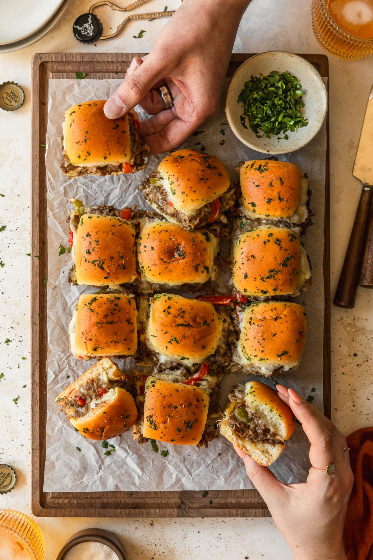 Hands reaching for Philly cheesesteak sliders on a wood board next to glasses of beer, white plates, and a beer opener with a tan background.