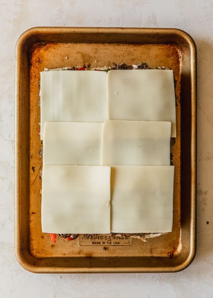 Philly cheesesteak sliders topped with American cheese on a gold sheet pan.