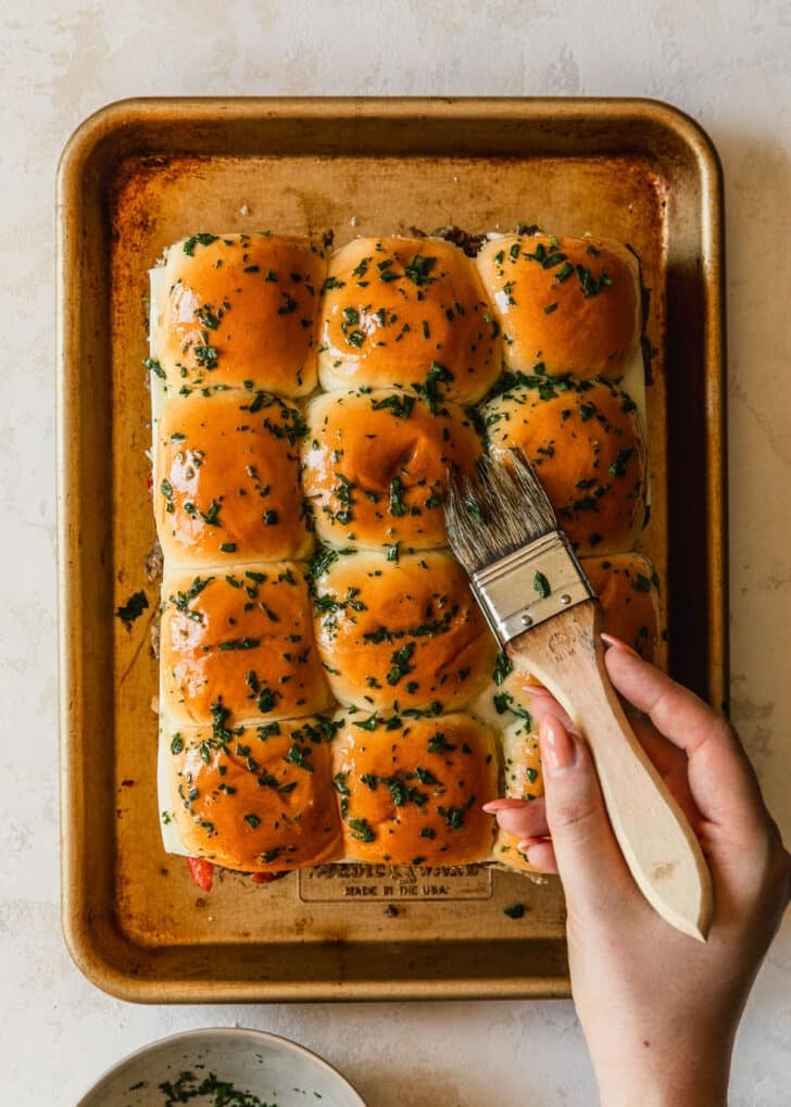 A hand brushing parsley butter over rolls on a gold sheet pan.